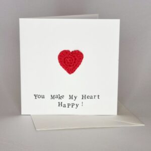 A square white greetings card with a red heart on the front with the words 'you make my heart happy'. a white envelope and grey white background