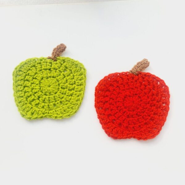 Crochet apple coasters by Sarah Lou Crafts. One green and one red, each with a small brown stem at the top, laying side by side on a white table.