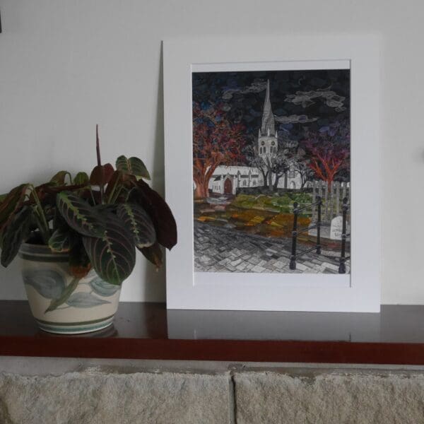 Picture of nighttime scene of floodlit church with surrounding winter trees and paving, shown in mount on shelf with pot plant beside.