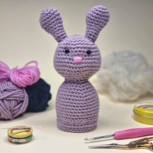 A lilac crochet bunny rabbit surrounded by crochet tools, yarn and wool stuffing.