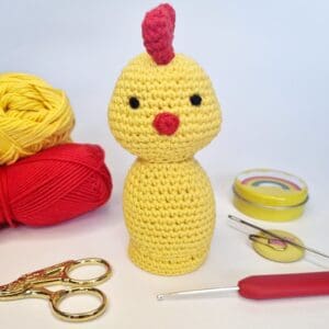A crochet yellow chicken with yarn and crochet tools scattered about on a plain white background