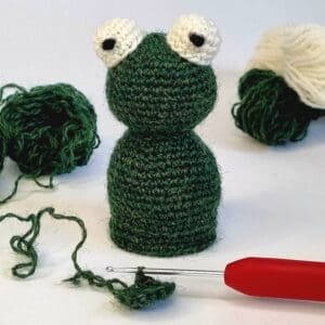 a green crochet frog sat next to a red crochet hook and some green and cream balls of wool