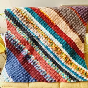 Crochet lap blanket by Sarah Lou Crafts using mixed aran yarn in a C2C pattern to create a multicoloured diagonal striped pattern.