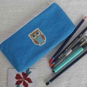 Sky blue zipped pencil case with cartoon owl stitched onto centre of front, shown with selection of pens and pencils