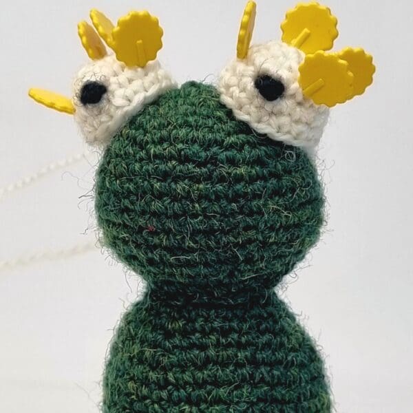 a close up head shot of a crochet frog in bottle green with yellow headed pins keeping crochet eyes in place