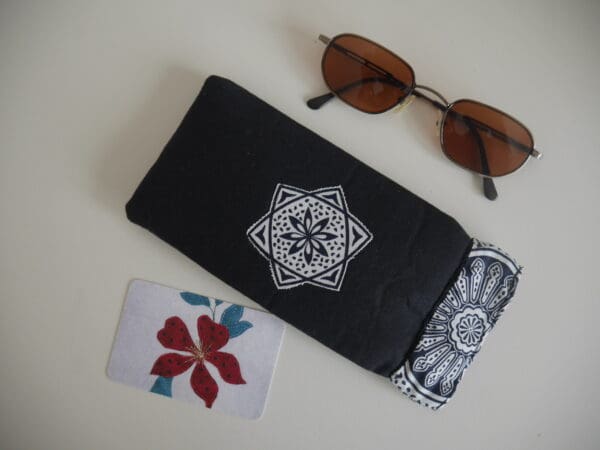 Black rectangular glasses case with black and white geometric design appliqued onto centre and black and white printed trim around the opening. Shown with sunglasses.