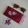Deep red rectangular glasses case with red and white stitched leaf design on centre and red and white print fabric cuff around the opening. Shown with sunglasses