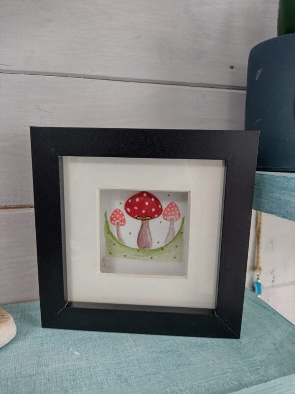 Three red and white toadstools hand painted in water colour paints, one of which is made up of a hand painted clam shell. Framed in a black box frame with mount.