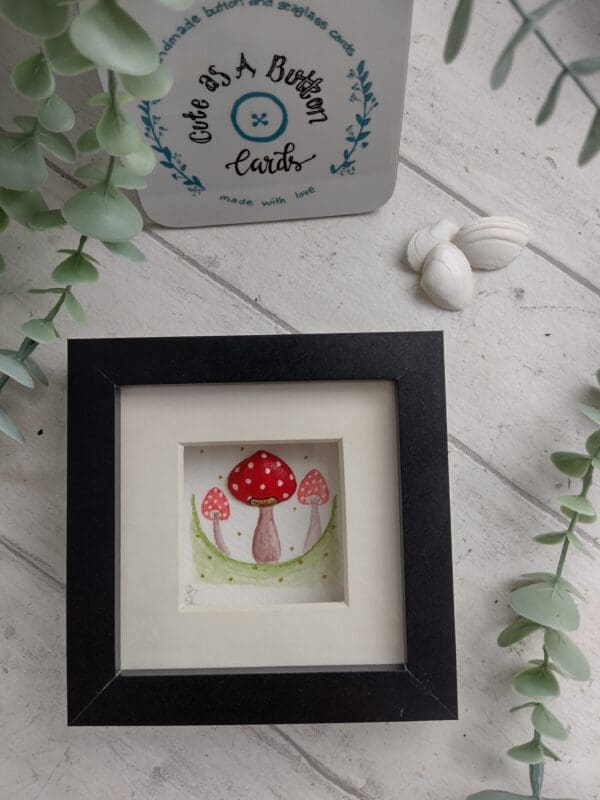 Three red and white toadstools hand painted in water colour paints, one of which is made up of a hand painted clam shell. Framed in a black box frame with mount.