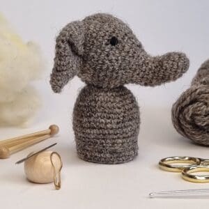 Crochet amigurumi elephant sat next to crochet accessories on a white background