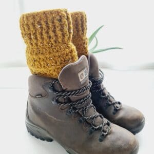 Mustard Yellow crochet boot cuffs made by Sarah Lou Crafts, shown in brown Scarpa Hiking Boots on a white table with green aloe vera plant in the back ground.