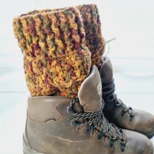 Crochet boot cuffs made by Sarah Lou Crafts, in a autumn russet colourway of red, yellow and green, shown in brown Scarpa Hiking Boots on a white table with green aloe vera plant in the back ground.