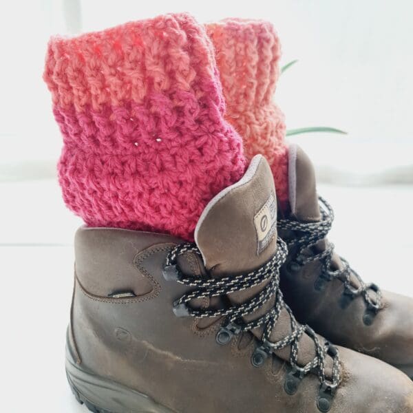 Crochet boot cuffs made by Sarah Lou Crafts, in shades of red and pink, shown in brown Scarpa Hiking Boots on a white table with green aloe vera plant in the back ground.