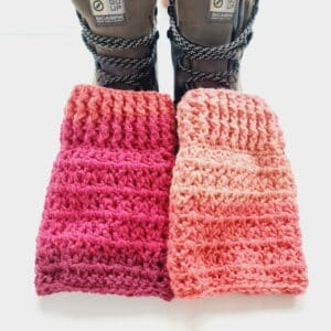 Crochet boot cuffs made by Sarah Lou Crafts, in shades of red and pink, shown in brown Scarpa Hiking Boots on a white table with green aloe vera plant in the back ground.