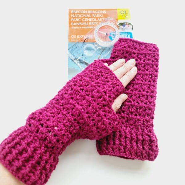 Plum, deep pink crochet fingerless mitts, made by Sarah Lou Crafts. Show on makers left hand resting on top of right mitts and OS map with a compass.