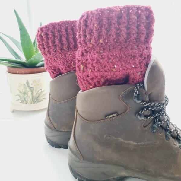 Crochet boot cuffs made by Sarah Lou Crafts, in a red colourway called Berry. Shown in brown Scarpa Hiking Boots on a white table with green aloe vera plant in the back ground.