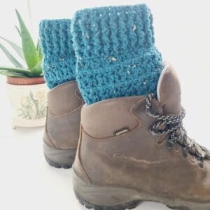 Crochet boot cuffs made by Sarah Lou Crafts, in a blue colourway called Ocean. Shown in brown Scarpa Hiking Boots on a white table with green aloe vera plant in the back ground.