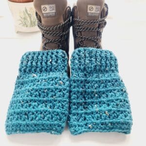 Crochet boot cuffs made by Sarah Lou Crafts, in a blue colourway called Ocean. Shown in brown Scarpa Hiking Boots on a white table with green aloe vera plant in the back ground.