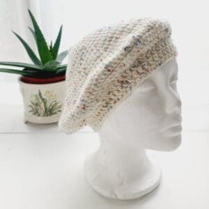 Crochet winter beret made by Sarah Lou Crafts using premium acrylic yarn in a cream colour with multicoloured flecks throughout, called Nougat. Shown on white mannequin head with plaint in back ground.