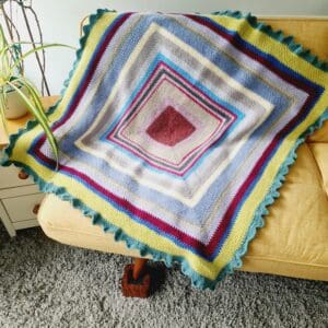 Crochet lap blanket by Sarah Lou Crafts using mixed DK yarns in a moss stitch in the round pattern to create a multicoloured striped pattern. Shown draped on a yellow sofa.