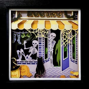 A 23cm shadow box featuring a spooky "Boo Store" with skeletons, a ghost, and a cat. The scene is intricately cut from card and has a black frame.