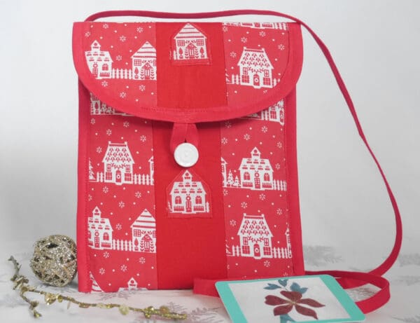 Small patchwork bag made from red and white cotton fabrics showing houses in snow, with red trim and narrow strap, white button closure