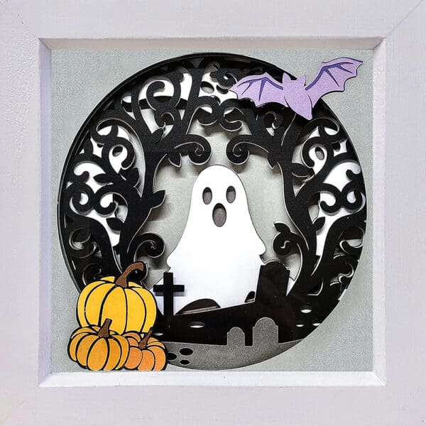 A 18cm shadow box featuring a ghost, a bat, pumpkins, and a graveyard. The scene is intricately cut from paper and framed in white.