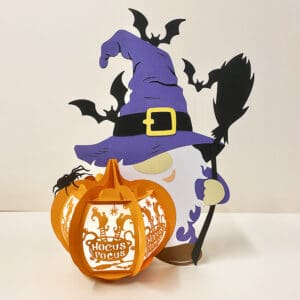 A 25cm Hocus Pocus gnome lantern featuring a gnome holding a broom and standing beside a pumpkin lantern with the Hocus Pocus phrase. Bats fly above, and the lantern is illuminated by battery-powered fairy lights.