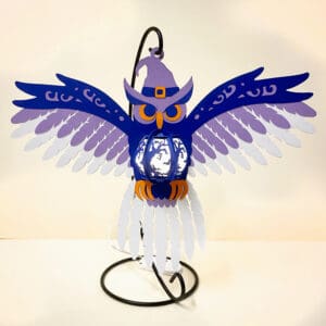 A 25cm owl lantern made from cardstock. The owl is wearing a witch's hat and holding a pumpkin lantern with jack-o'-lantern faces. The lantern is illuminated by fairy lights, and the owl is standing on a metal stand.