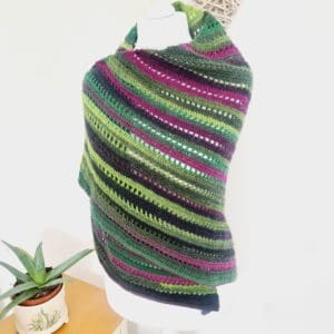 Crochet shawl wrap by Sarah Lou Crafts in a variegated yarn called Rocket of purple and green shades, shown on a white dress makers mannequin.