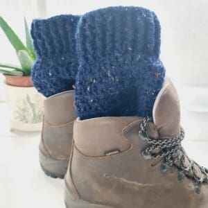 Crochet boot cuffs made by Sarah Lou Crafts, in a navy blue colourway with multicoloured flecks. Shown in brown Scarpa Hiking Boots on a white table with green aloe vera plant in the back ground.