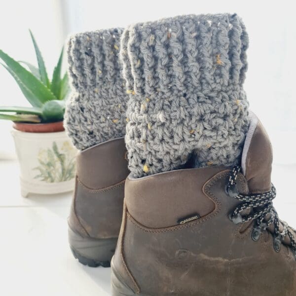 Crochet boot cuffs made by Sarah Lou Crafts, in a soft sage green colourway with multicoloured flecks. Shown in brown Scarpa Hiking Boots on a white table with green aloe vera plant in the back ground.