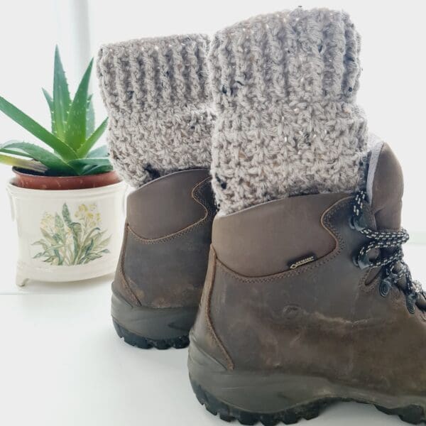 Crochet boot cuffs made by Sarah Lou Crafts, in a soft cream colourway called Starling, with black and cream flecks throughout. Shown in brown Scarpa Hiking Boots on a white table with green aloe vera plant in the back ground.