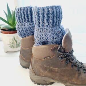 Crochet boot cuffs made by Sarah Lou Crafts, in a soft blue colourway called Stormy, with brown and cream flecks. Shown in brown Scarpa Hiking Boots on a white table with green aloe vera plant in the back ground.