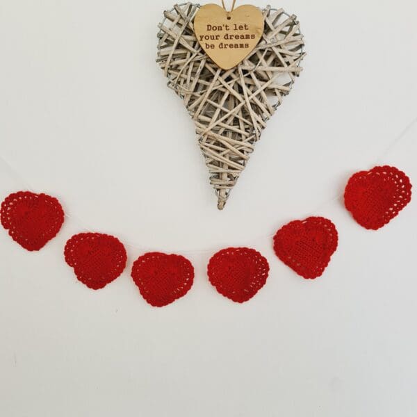 Crochet red heart garland set of 6 made by Sarah Lou Crafts shown strung on a white wall with a wicker heart hanging decoration.