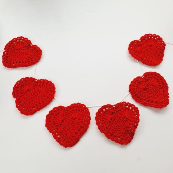 Six red crochet hearts by Sarah Lou Crafts shown hung on string and arranged in a U shape laying flat on a white table.