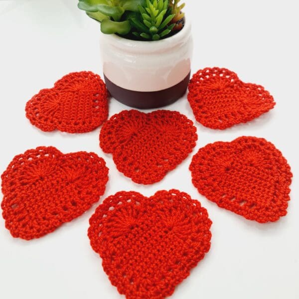 Set of 6 red heart crochet coaster mats made by Sarah Lou Crafts shown arranged on a white table with a small decorative house plant.