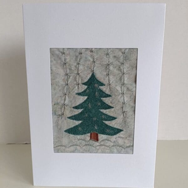 Greetings card showing fabric tree on grey fabric background with machine embroidered snowflakes.