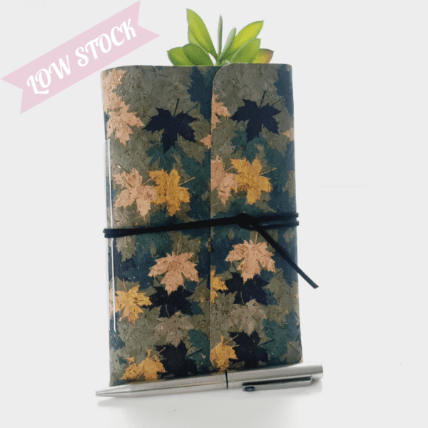 Cork fabric notebook journal with green and gold leaves pattern. Photographed standing again a green plant with silver pen in front.