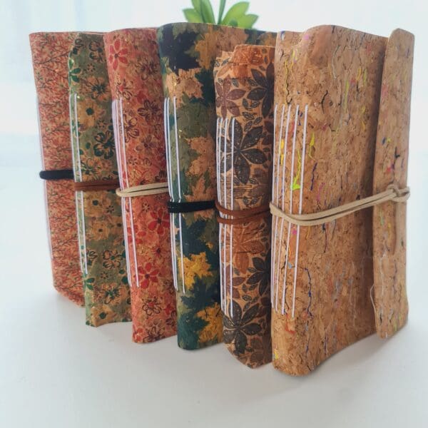 Group photo of 6 cork fabric notebook journals made by Sarah Lou Crafts; Pink Blossom, Green Floral, Pink Floral, Green Leaves, Flower Print, and Rainbow Fleck.