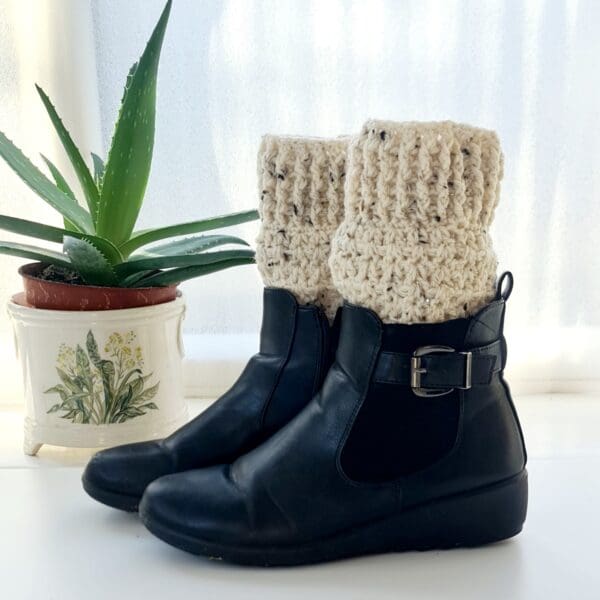 Cream berber crochet boot cuffs made by Sarah Lou Crafts, shown long in black ankle boots on a white table with green aloe vera plant in the background.