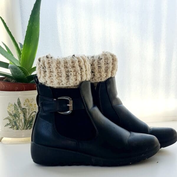 Cream cochet boot cuffs made by Sarah Lou Crafts. Shown in black ankle boots on a white table with green aloe vera plant in the background.