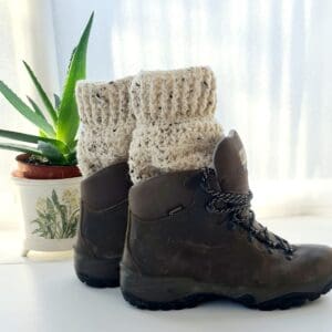 Crochet boot cuffs made by Sarah Lou Crafts, in a cream yarn with black flecks throughout. Shown in brown Scarpa Hiking Boots on a white table with green aloe vera plant in the back ground.