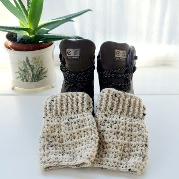 Crochet ankle warmer boot cuffs made by Sarah Lou Crafts, in Cream Berber yarn, shown laying on brown hiking boots on a white table with green aloe vera plant in the background.