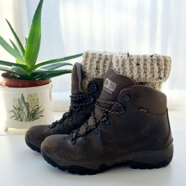 Crochet boot cuffs made by Sarah Lou Crafts, in a cream colourway called Berber. Shown in black ankle boots on a white table with green aloe vera plant in the background.