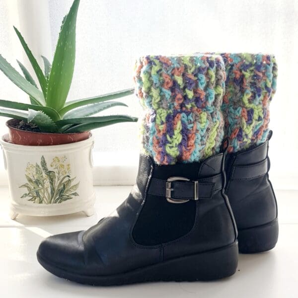 Multicoloured Fireworks crochet boot cuffs made by Sarah Lou Crafts, shown long in black ankle boots on a white table with green aloe vera plant in the background.