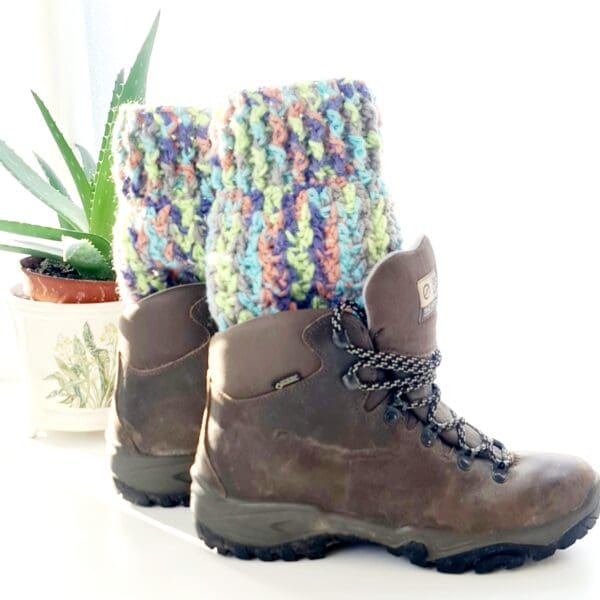 Crochet boot cuffs made by Sarah Lou Crafts, in a multicoloured yarn called Fireworks. Shown in brown Scarpa Hiking Boots on a white table with green aloe vera plant in the back ground.