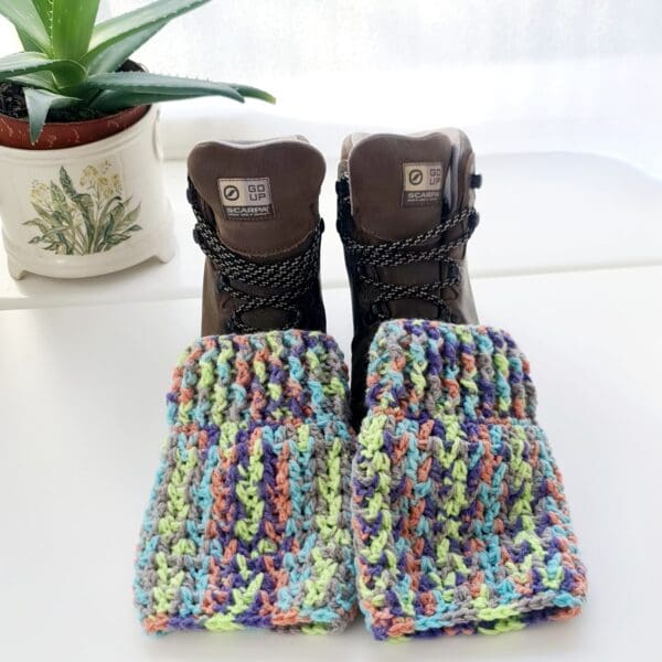 Crochet ankle warmer boot cuffs made by Sarah Lou Crafts, in Fireworks coloured yarn, shown laying on brown hiking boots on a white table with green aloe vera plant in the background.