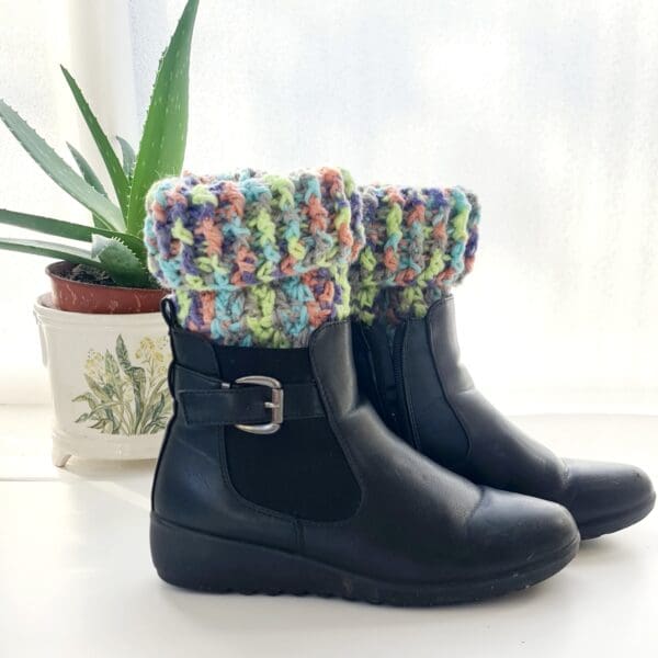 Multicoloured crochet boot cuffs made by Sarah Lou Crafts in a colourway called Fireworks. Shown in black ankle boots on a white table with green aloe vera plant in the background.