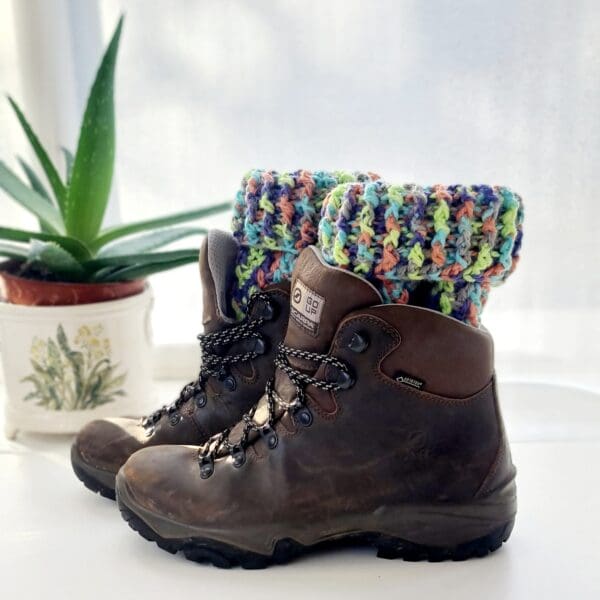 Crochet boot cuffs made by Sarah Lou Crafts, in a colourful yarn called Fireworks. Shown in black ankle boots on a white table with green aloe vera plant in the background.